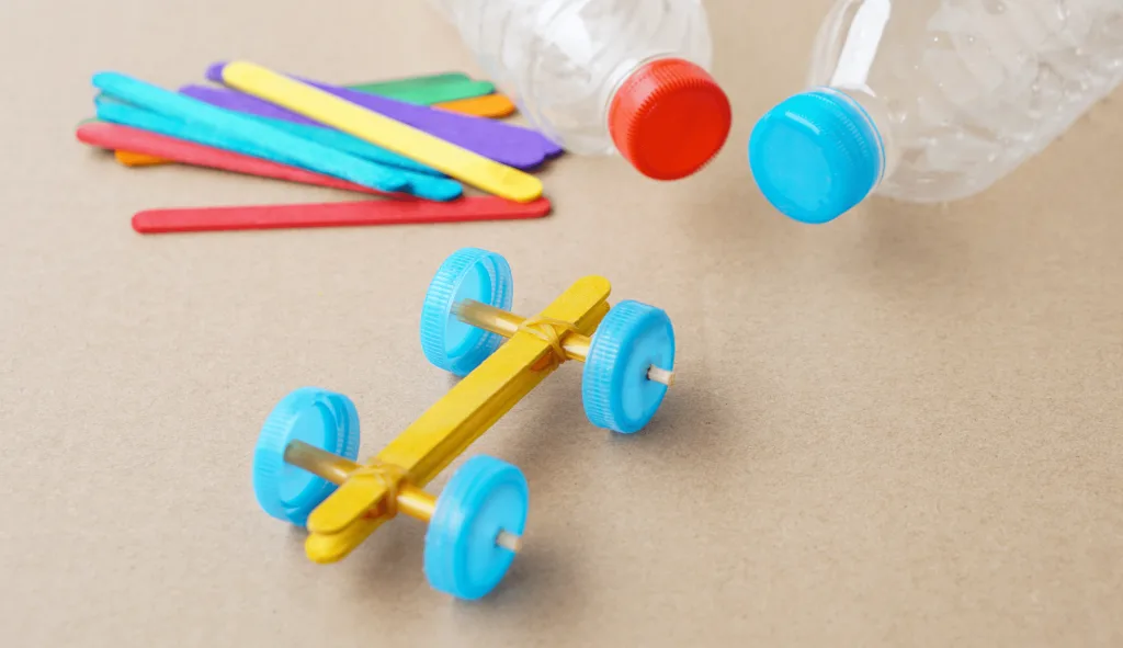 Mini carrinho de corrida feito com pregadores de roupa e botões coloridos sobre fundo de papel craft.