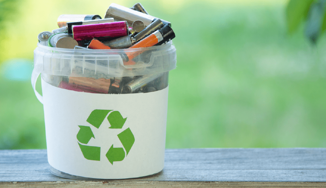 Balde de reciclagem cheio de baterias usadas sobre um fundo verde, simbolizando o descarte responsável e a importância de reciclar baterias.