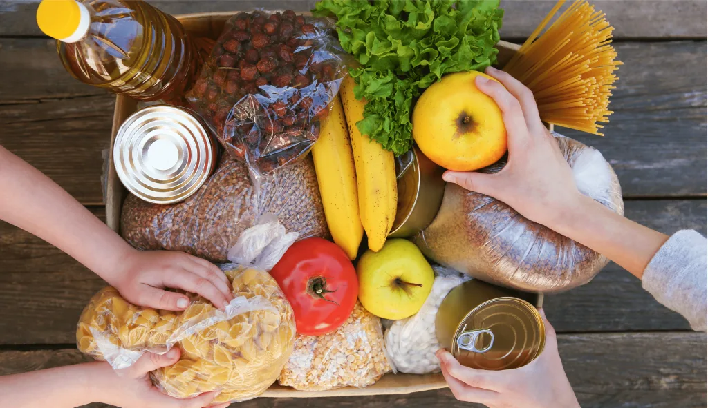 Mãos organizando uma caixa de doação com alimentos diversos, representando a solidariedade no combate à fome e desperdício de alimentos.